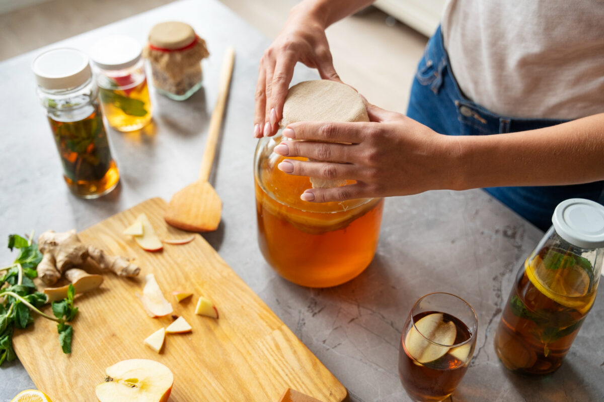 Close-up of yellow nails soaking in a bowl of apple cider vinegar solution with lemon slices, surrounded by cotton pads and a small bottle of ACV on a wooden surface.