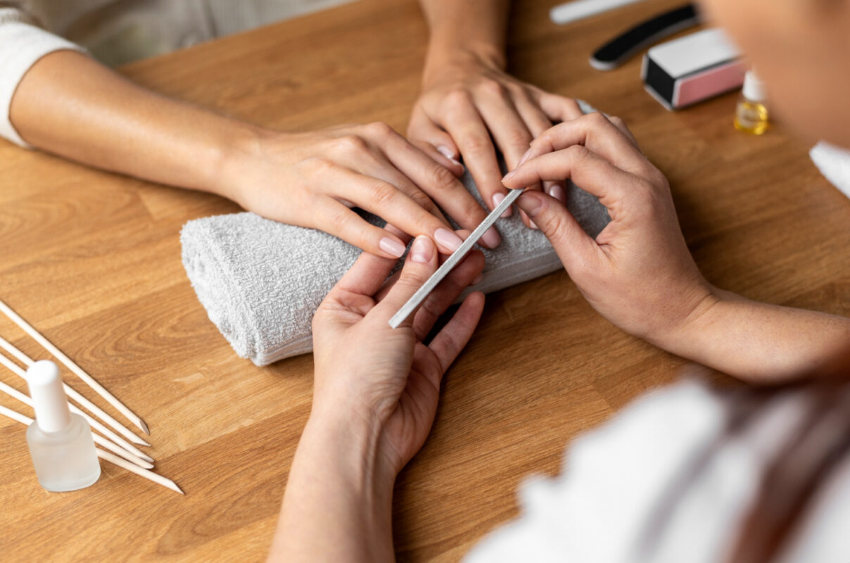 A nail technician filing a client's nails during a manicure session, with tools and nail products on a wooden table, showcasing a gentle and hypoallergenic nail treatment.